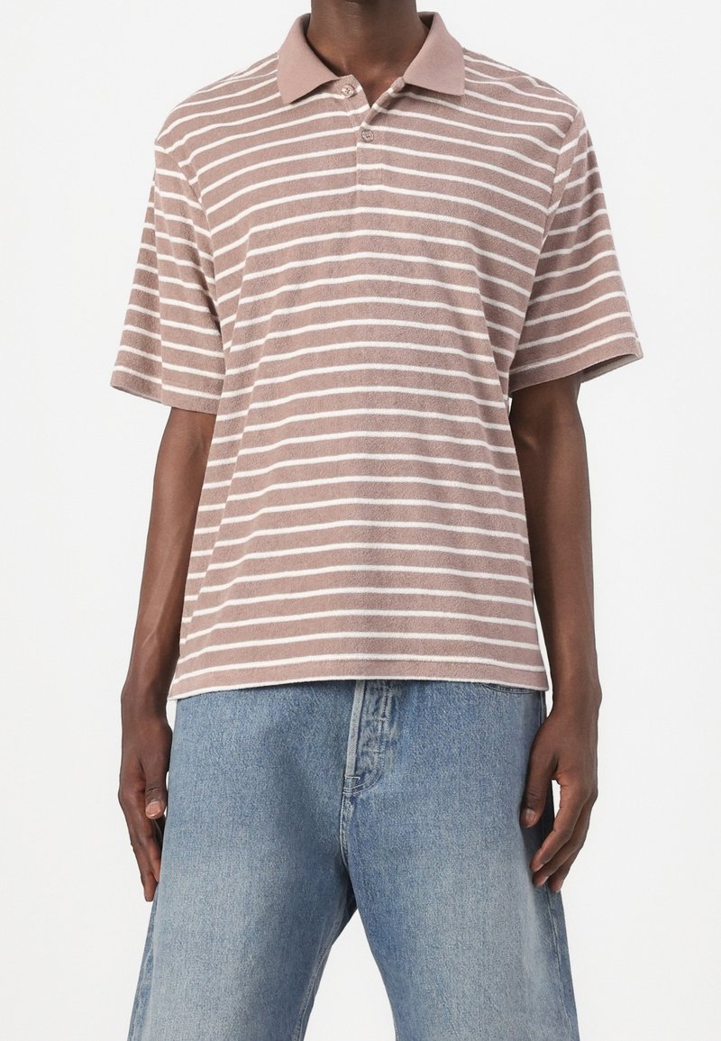 Personne portant un polo à manches courtes marron clair avec des rayures horizontales blanches et un jean en denim bleu clair.