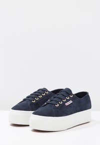 Navy suede platform sneakers with gold eyelets, white textured rubber sole, and a small brand tag on the side.