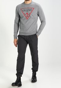 Hombre con un sudadera de manga larga gris con el logo rojo de Guess, pantalones de chándal gris oscuro y botas negras de tobillo, de pie con una mano en el bolsillo.