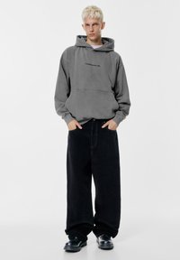 Szary hoodie z kapturem i kieszenią z przodu, z małym nadrukiem tekstowym. W zestawieniu z luźnymi czarnymi spodniami z kordurowej tkaniny i czarnymi butami.