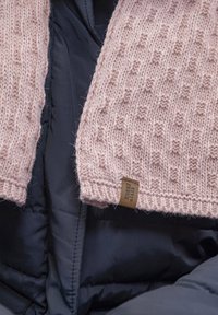 Sciarpa rosa lavorata a maglia con un motivo testurizzato, dotata di un'etichetta marrone. È sovrapposta a un giubbotto imbottito nero. Materiale morbido e caldo.