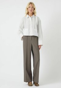 Camicia corta bianca con colletto e due tasche frontali; abbinata a pantaloni marroni a vita alta e gamba larga. Tessuto morbido e colori neutri.