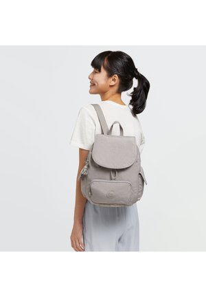 Sac à dos en tissu gris clair avec un rabat, poche avant, poches latérales et bretelles ajustables. Présente un logo et une surface texturée.