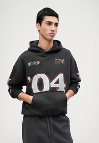 Melnā kapuce ar priekšējo kabatu, lielu sudraba '04' druku un logo, ieskaitot Oracle un Red Bull Racing. Mīksts audums, ikdienišķa piegriezuma.