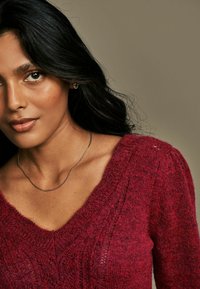 Maglione a V in punta, lavorato a maglia di un profondo rosso, che mette in mostra una texture a coste e dettagli intricati del motivo. Include una delicata collana d'argento.
