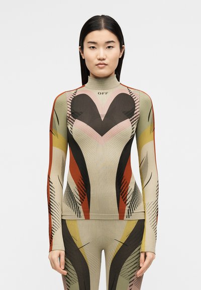 Γυναίκα με ίσια μαύρα μαλλιά, που φοράει εφαρμοστό, πολύχρωμο γεωμετρικό πουλόβερ με ζιβάγκο και ταιριαστό παντελόνι, στέκεται μπροστά σε λευκό φόντο.