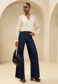 Light yellow, textured blouse with ruffled cuffs; dark blue wide-leg jeans; black handbag; platform heels with wood accent.