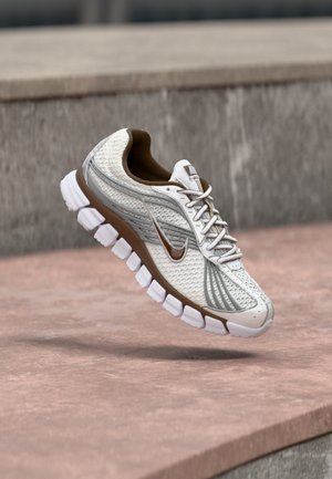 Άσπρο και καφέ παπούτσι για τρέξιμο Nike με επάνω μέρος από δίχτυ και τμηματική σόλα, αιωρούμενο πάνω από τσιμεντένια σκαλοπάτια σε εξωτερικό χώρο.