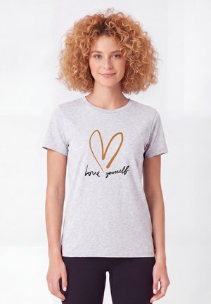 Frau mit lockigem Haar, die ein hellgraues T-Shirt mit einem orangefarbenen Herz und den Worten "Liebe dich selbst" in schwarzer Schrift trägt.
