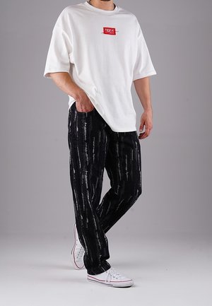 Mężczyzna noszący za dużą białą koszulkę z czerwonym logo, czarne spodnie w wzory oraz białe trampki na jednolitym szarym tle.