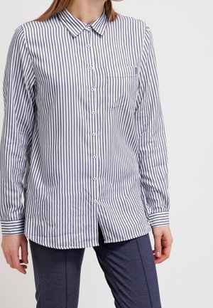 Kvinde iført en hvid langærmet skjorte med tynde lodrette grå striber og mørkeblå bukser, stående mod en ensfarvet lys baggrund.