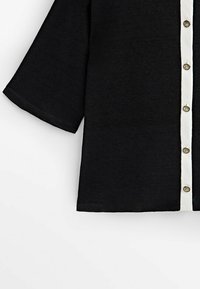 Chemise noire à manches courtes avec une patte de boutonnage blanche ornée de boutons ronds dorés, confectionnée dans un tissu doux et texturé.