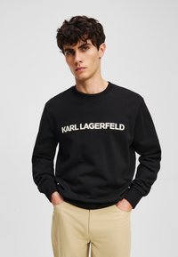 Fekete pulóver kerek nyakkivágással, a mellkason fehér "KARL LAGERFELD" felirattal; világos bézs nadrággal párosítva.