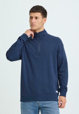 Jonge man die de kraag van een marineblauwe sweater met halve rits rechtzet, draagt lichtblauwe jeans, staand tegen een vlakke lichtgrijze achtergrond.