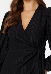 Vestido negro ajustado con tejido texturizado que presenta rayas verticales, escote en V profundo y mangas largas abullonadas, asegurado con un cinturón atado.