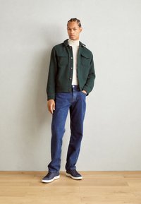 Dark green jacket with two front pockets, button closure, white turtleneck, blue jeans, and dark sneakers on wooden floor against gray wall.