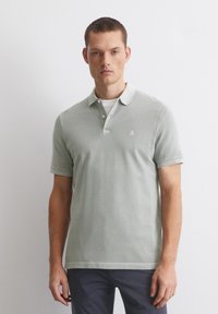 Mężczyzna w jasnoszarej koszulce polo z krótkim rękawem, w białej koszulce pod spodem i ciemnych spodniach, stoi na tle jednolitej białej ściany.