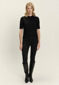 Maglione nero a maniche corte con un design aderente, lavorazione a maglia testurizzata e tre bottoni decorativi sulla spalla. Abbinato a jeans skinny neri e stivali al ginocchio.