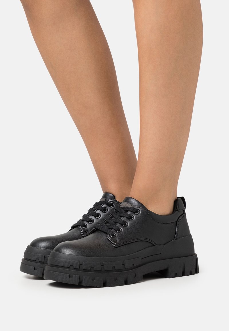 Personne portant des chaussures en cuir noires à lacets, robustes, avec des semelles épaisses et texturées, sur un fond clair uni.