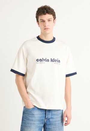 Short-sleeve cream t-shirt with navy blue ribbed collar and cuffs, featuring bold "calvin klein" text and city details printed on the front.