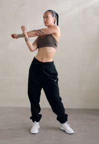 Sujetador deportivo marrón con el logo de Nike, pantalones deportivos negros con puños y zapatillas deportivas blancas. Persona en una pose de estiramiento, con tatuajes visibles.