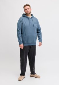 Μπλε φούτερ με κουκούλα, μπροστινή τσέπη και κορδόνι, με διακριτικό λογότυπο. Συνδυασμένο με σκούρο παντελόνι και ανοιχτόχρωμα παπούτσια.