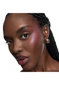 Gros plan sur un visage aux cheveux tressés, mettant en valeur une peau foncée, un sourcil saillant, des lèvres bordeaux et des boucles d'oreilles circulaires dorées.