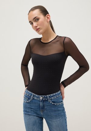 Mujer joven con una blusa negra transparente de manga larga con pequeños remaches y vaqueros azules con un cinturón decorado con perlas, de pie con las manos en las caderas.