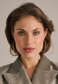 Modell som bär en strukturerad, ljusguldblazer med spetsiga slag, guldörhängen i form av ringar och ett delikat guldhalsband. Naturlig sminkning och vågigt hår.