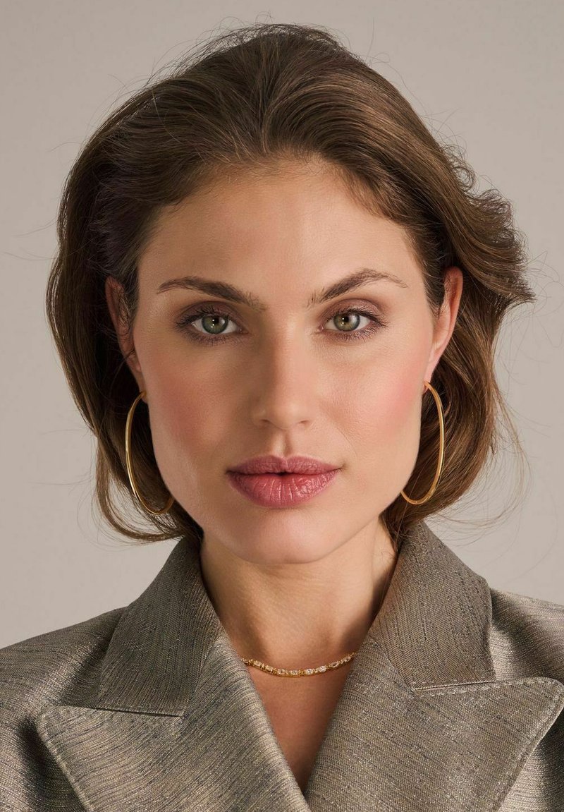 Modell som bär en strukturerad, ljusguldblazer med spetsiga slag, guldörhängen i form av ringar och ett delikat guldhalsband. Naturlig sminkning och vågigt hår.