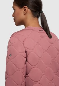 Veste rose matelassée avec une texture en vagues, épaules arrondies et détail de logo sur la manche. Matériau synthétique lisse.