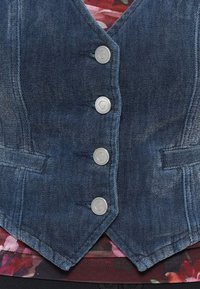 Denim veste ar pieguļošu dizainu, dziļi zilu krāsu, piecām sudraba pogām un divām priekšējām kabatām. Zemē redzams ziedu raksts.