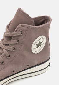 Pruunist nubukist kõrge ülemise osaga tossud, mis on seotud nööridega, valge kummist tald ja ümmargune "Converse All Star" märk külje peal.