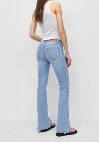 Jean évasé bleu clair avec une finition délavée, cinq poches et un patch en cuir sur la ceinture arrière. Associé à un débardeur blanc.