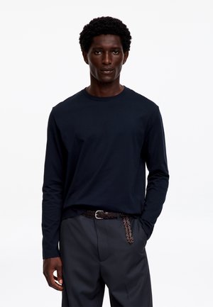 Homme à la peau foncée et aux cheveux bouclés portant une chemise noire à manches longues, un pantalon foncé et une ceinture tressée marron, debout devant un fond blanc.