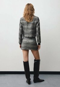 Czarnobiały tweedowy krótki żakiet i mini spódnica, połączone z czarnymi, błyszczącymi kozakami do kolan, widziane od tyłu na neutralnym tle.