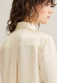 Kvinde med krøllet brunt hår iført en cremefarvet langærmet skjorte, vist fra skulderen bagfra og i profil mod en ensfarvet baggrund.