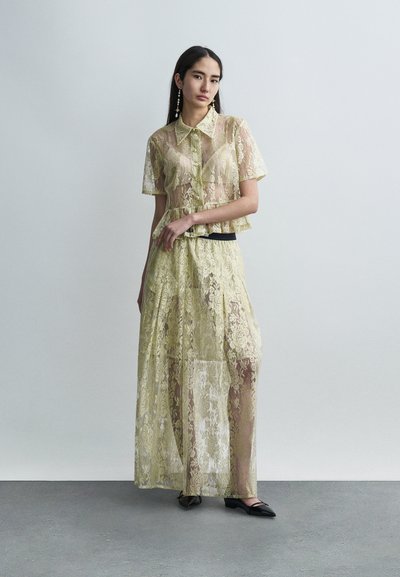 Σετ δύο τεμαχίων από δαντέλα σε ανοιχτό κίτρινο χρώμα, που περιλαμβάνει ένα κοντομάνικο πουκάμισο με γιακά και μια μακριά, ρέουσα φούστα, και τα δύο με φυτικό μοτίβο.