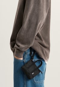 Persoon die een blauwe spijkerbroek en een bruin shirt met lange mouwen draagt, met een klein zwart Vans tasje dat aan de broekzak van de jeans geklemd is.