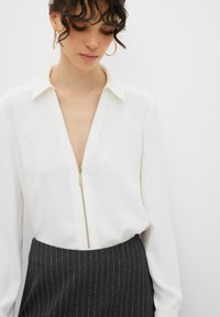Blusa bianca a maniche lunghe con zip davanti e colletto, abbinata a una gonna scura a righe. Tessuto liscio e design aderente.