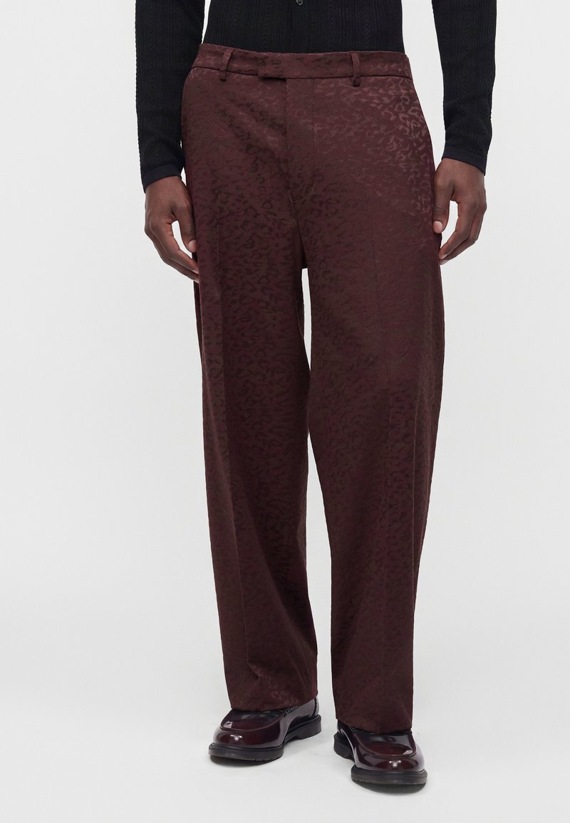 Homme portant un pantalon large brun rouge foncé avec un motif subtil, un haut noir à manches longues et des chaussures habillées marron brillantes, debout les mains le long du corps.