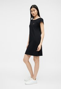 Νεαρή γυναίκα στέκεται, φορώντας κοντομάνικο μαύρο φόρεμα με μικρό λογότυπο στο στήθος και άσπρα αθλητικά παπούτσια, μπροστά σε απλό φωτεινό φόντο.