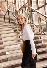 Chemise blanche à manches longues, pantalon noir, tenant des dossiers. Le modèle se tient sur un escalier avec une rampe en métal et de grandes fenêtres en arrière-plan.