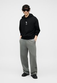 Άνδρας με μαύρη μπλούζα με κουκούλα, γκρι παντελόνια με φαρδιά μπατζάκια και λογότυπο κοντά στον αστράγαλο, μαύρα αθλητικά παπούτσια και μεγάλα μαύρα γυαλιά ηλίου, στέκεται με τα χέρια στις τσέπες.