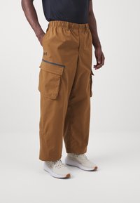 Hnědé cargo kalhoty s elastickým pasem, které mají několik kapes a kontrastní lemování. Nosit s světle šedými sportovními botami.