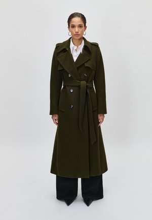 Trench-coat double boutonnage olive avec une taille ceinturée, des épaulettes et un large col, porté par-dessus un pantalon large sombre.