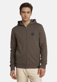 Sudadera con capucha y cremallera en verde olivo, cuenta con un parche de logotipo en navy en el pecho, bolsillos laterales y puños y dobladillo acanalados. Textura de tela suave.