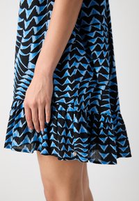 Abito nero con motivo a zigzag blu; presenta un volant all'orlo e un bottone decorativo visibile vicino al fondo. Tessuto leggero.