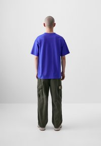 T-shirt violet à manches courtes en coton, associé à un pantalon cargo vert olive avec des poches latérales et une patch sur la jambe droite.