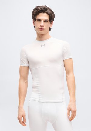 Mlajši moški s kratkimi rjavimi lasmi, oblečen v tesno belo športno majico in ujemajoče stisnjene hlače, stoji pred enostavnim svetlim ozadjem.